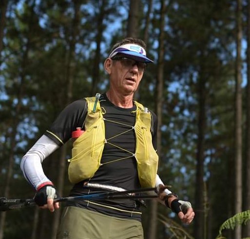 Fred Richardson running with poles through pine forest, race vest on, looking up the trail.
