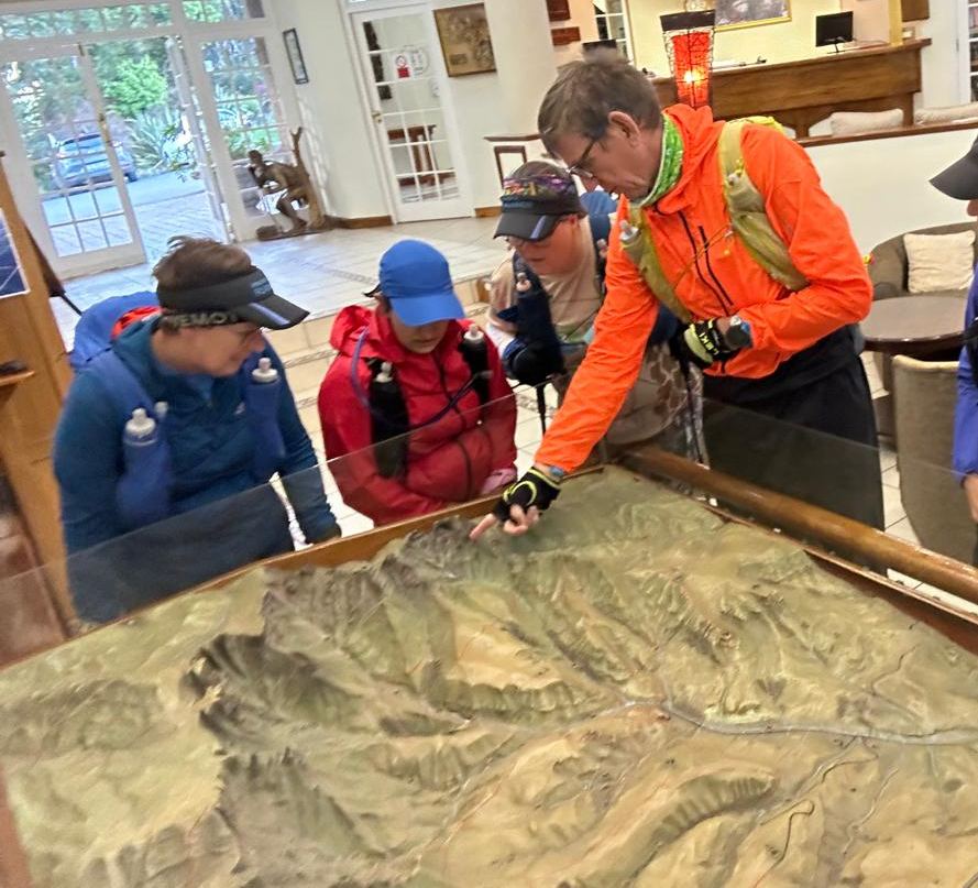 A coach points at a ridge on a large relief model of the escarpment while four trail runners in kit lean in to study the terrain.